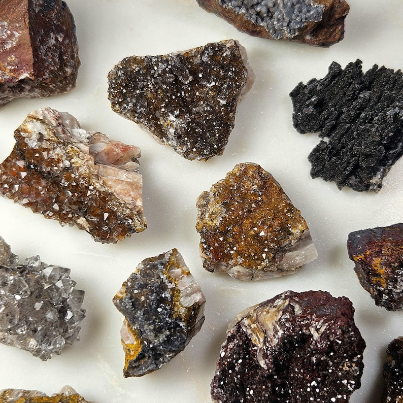 May include: A collection of various raw mineral specimens, including quartz and other crystalline formations. The stones display a range of colors, from deep browns and blacks to lighter tans and grays, with sparkling crystal inclusions.