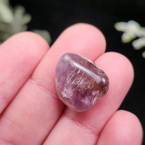 May include: A polished, tumbled amethyst gemstone, displaying a mix of purple and brown hues. The stone is smooth and rounded, held in a person's hand, with a natural, organic appearance. The gemstone is a small, decorative item.