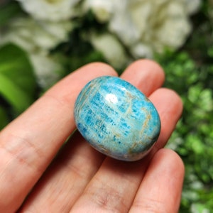 May include: A polished, oval-shaped gemstone in shades of turquoise and blue, with subtle brown veining. The stone is held in a hand, showcasing its smooth, rounded form. The background is blurred, with hints of green and white.