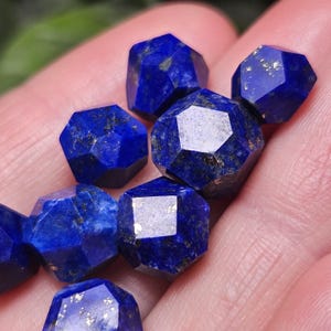 May include: A collection of faceted lapis lazuli gemstones, each displaying a deep royal blue color with natural golden pyrite inclusions. These small, multi-sided stones feature polished surfaces that reflect light across their geometric edges.