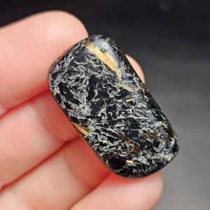 May include: A polished, rectangular gemstone with a black base color and intricate white and gold veining patterns. The stone is smooth and rounded, with a glossy finish. The overall appearance is dark and textured, with a natural, earthy aesthetic.