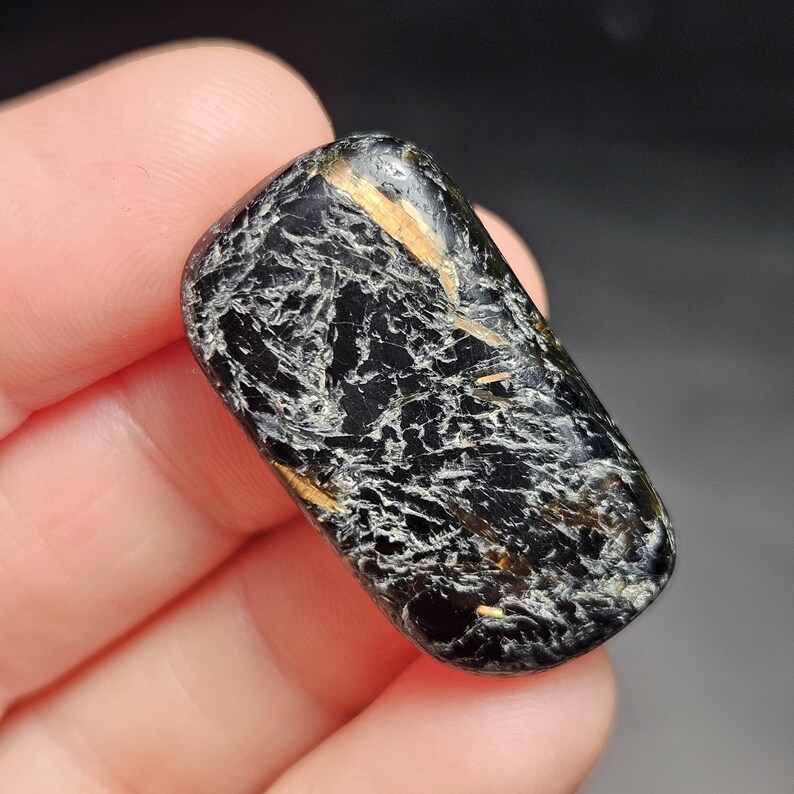 May include: A polished, rectangular gemstone with a black base color and intricate white and gold veining patterns. The stone is smooth and rounded, with a glossy finish. The overall appearance is dark and textured, with a natural, earthy aesthetic.