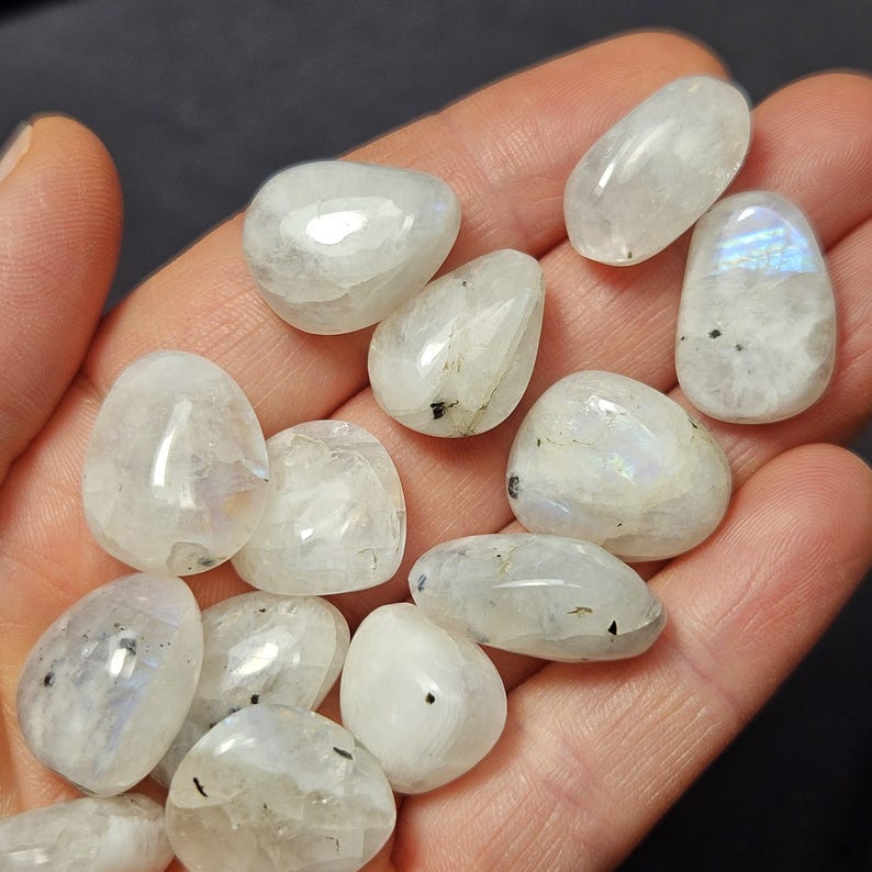 May include: A selection of polished moonstone gemstones. The stones are mainly white with iridescent flashes of blue and other colours. Each stone has a smooth, rounded shape and varying sizes. Some stones have small black inclusions.