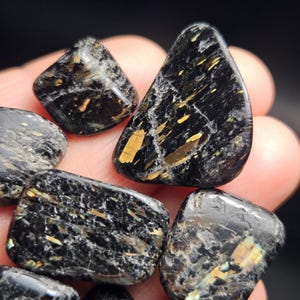 May include: A collection of polished black stones with gold-colored inclusions. The stones vary in shape, with some being rounded and others more angular. The gold inclusions create a striking contrast against the dark background.
