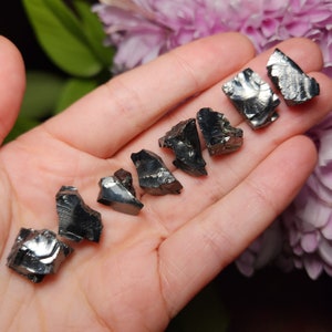 May include: A collection of eight black obsidian crystals, each with a unique shape and texture. The crystals are arranged in a row on a hand, showcasing their natural beauty.