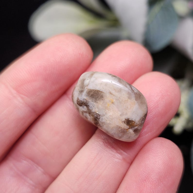 May include: A polished, oval-shaped gemstone with a mix of beige, brown, and translucent areas. The stone is smooth and appears to be held in a person's hand, with a blurred background of greenery.