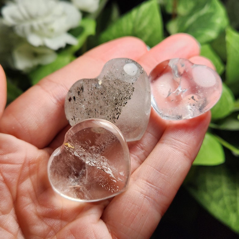 May include: Three heart-shaped clear quartz crystals, two are clear and one has inclusions of black and grey. The crystals are in the palm of a hand.
