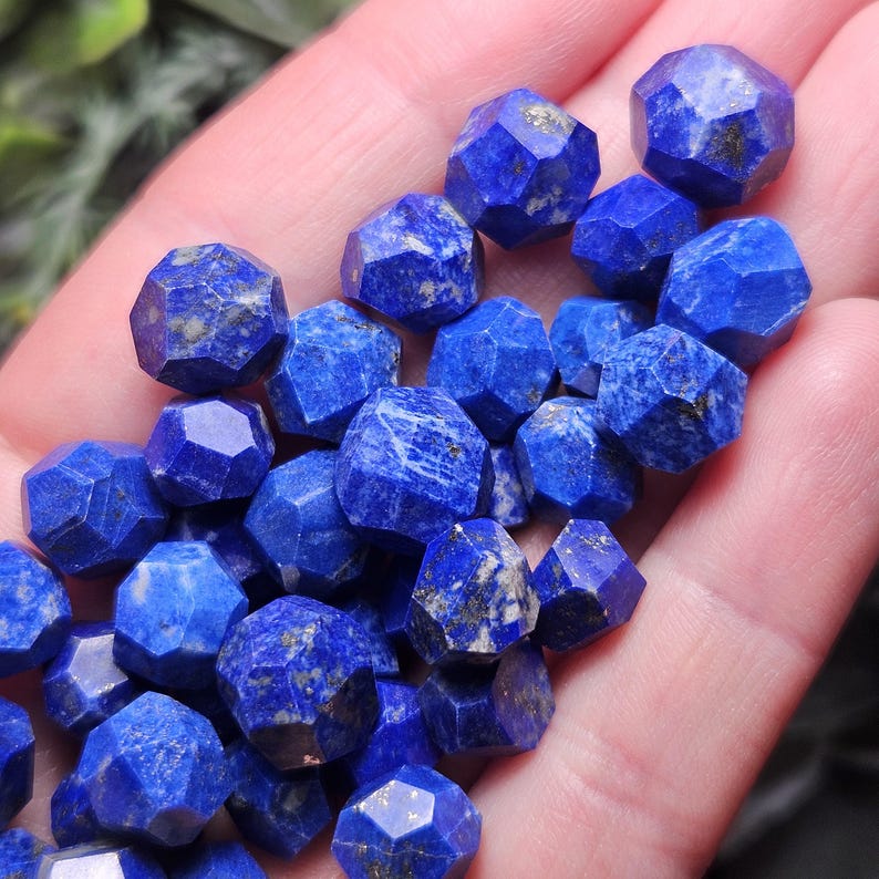 May include: A collection of small, faceted lapis lazuli beads with a deep blue color and natural gold-colored pyrite inclusions. Each bead features a multi-faceted geometric shape, creating a textured appearance across the group of loose stones.