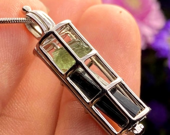May include: A silver-toned metal cage pendant featuring a rectangular frame that holds a small, translucent light green crystal at the top and a long, opaque black crystal at the bottom, suspended from a thin silver-toned metal chain necklace.