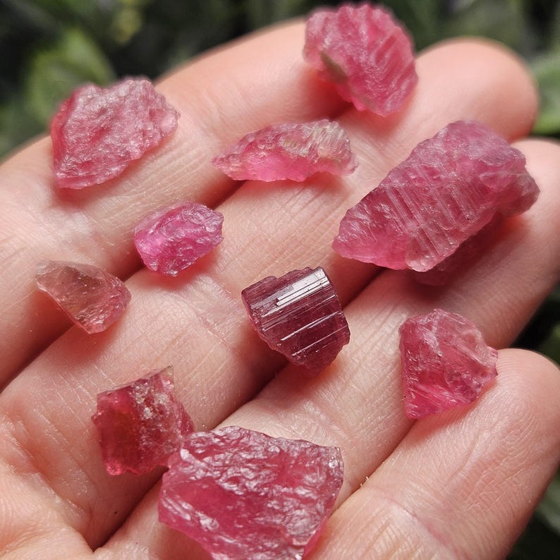 May include: A collection of small, raw pink tourmaline crystals with varying shapes and textures. The stones exhibit a vibrant, translucent to opaque magenta hue, with one central piece displaying distinct vertical striations on its surface.