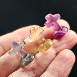 May include: Three small, colorful crystal crosses in shades of purple, yellow, and clear. Each cross is held in a hand, showcasing the unique color variations and translucent quality of the gemstones. The crosses have a rounded base.