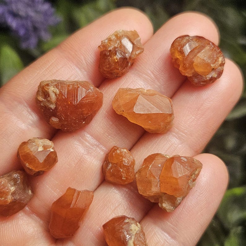 May include: A collection of raw, orange-brown garnet crystals, each with unique shapes and facets. The gemstones vary in size and are held in a hand, showcasing their natural, earthy tones and crystalline structures. The crystals are illuminated by natural light.