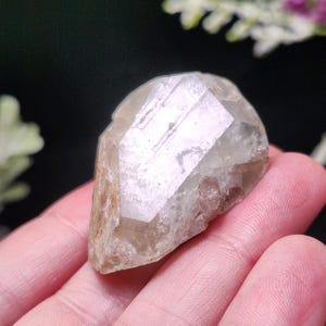 May include: A clear, faceted crystal with a light brown hue. The crystal is held in a hand, showcasing its natural shape and texture. The crystal's surface reflects light, highlighting its geometric structure. A natural gemstone.