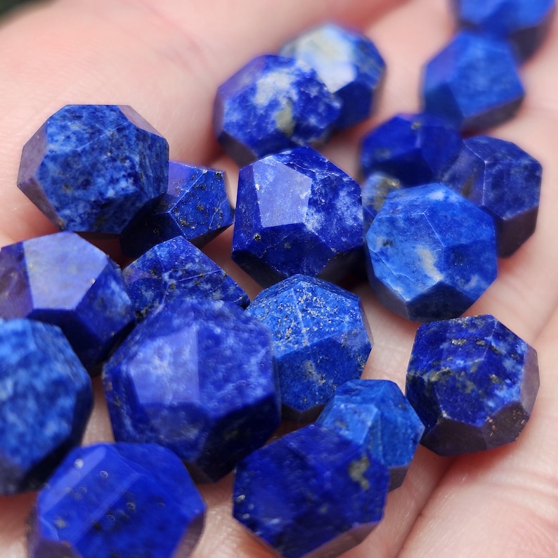 May include: A collection of small, faceted lapis lazuli gemstones with a deep blue color, featuring natural white calcite inclusions and golden pyrite flecks. Each stone is cut into a multi-sided geometric shape with sharp, polished edges.