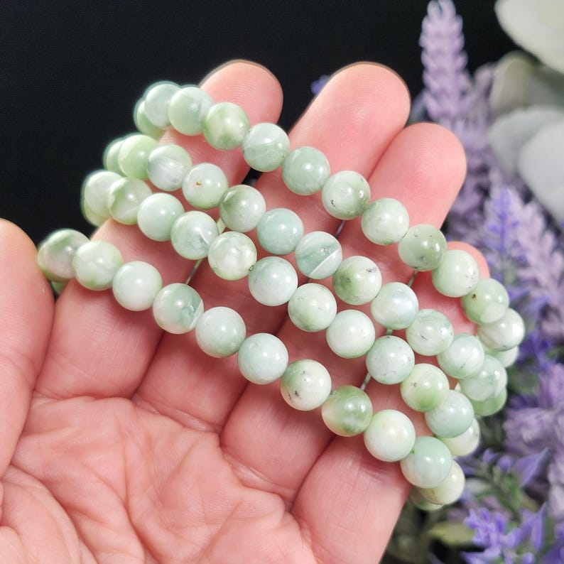 Peut inclure: Quatre bracelets de perles vert clair sont tenus dans une main. Les perles rondes ont un aspect marbr&eacute; avec des variations blanches et vert plus fonc&eacute;. L'arri&egrave;re-plan est flou, avec des touches de fleurs violettes et un fond noir.