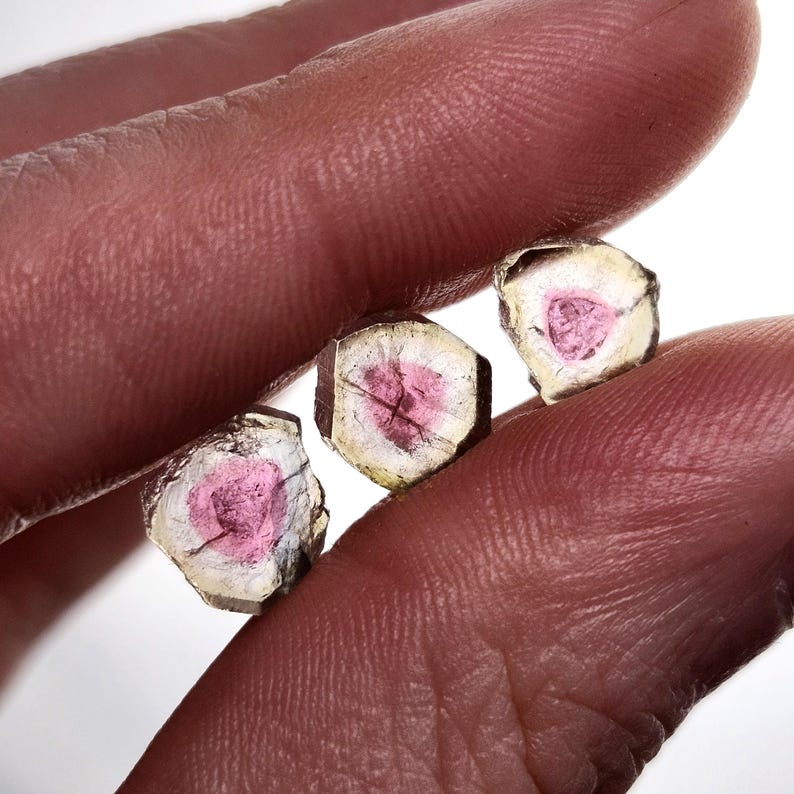 May include: Three small, raw watermelon tourmaline crystal slices featuring a vibrant pink centre surrounded by a translucent, pale green outer ring. Each piece has a distinct hexagonal shape with natural, rough edges and visible internal fractures.