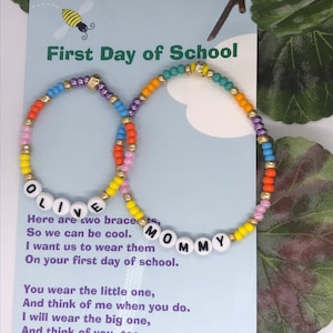 May include: Two colorful beaded bracelets with the words "LIVE" and "MOMMY" spelled out. The bracelets are on a card with the text "First Day of School" and a poem about the bracelets.