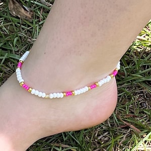 May include: White, pink, and gold beaded ankle bracelet. The bracelet is made of small beads and is worn on a person's ankle.