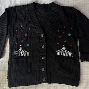 May include: A black cable-knit cardigan featuring a V-neckline, long sleeves with ribbed cuffs, and a button-front closure. The front panels are decorated with embroidered white circus tents and small, colourful stars in teal, purple, and red.