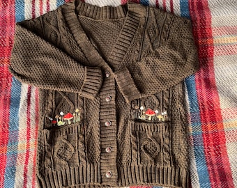 May include: A brown cable knit cardigan with a button closure and two pockets. The pockets are embroidered with red and white mushrooms.