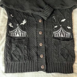 May include: Black knitted cardigan with a cable knit pattern and white embroidered details of ghosts, bats, and tents. The cardigan has a hood and a button closure.