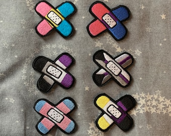 May include: Six embroidered iron-on patches shaped like band-aids with different color combinations. The patches are in the colors of the transgender, non-binary, bisexual, pansexual, lesbian, and gay pride flags.