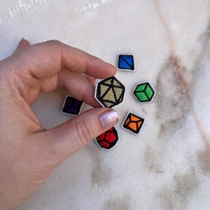 May include: Set of six embroidered iron-on patches featuring different coloured dice. The dice are in various shapes, including a cube, a pyramid, and a dodecahedron. The patches are perfect for adding a touch of whimsy to clothing, bags, or other accessories.