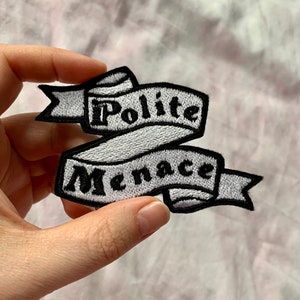 Può includere: Una toppa ricamata bianca con lettere nere che recitano "Polite Menace" su uno stendardo bianco.