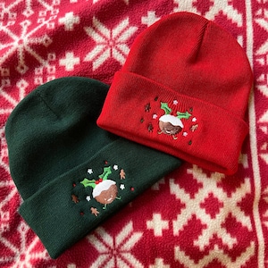 以下が含まれることがあります： 赤いニットのクリスマス帽子と緑のニットのクリスマス帽子が2つあり、クリスマスプディングの刺繍が施されています。帽子は赤と白の模様の背景の上に置かれています。