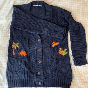 Peut inclure: Un cardigan en tricot bleu marine à câble avec deux poches plaquées et des motifs brodés de dinosaures et de palmiers.