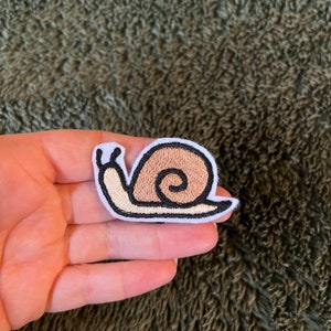 Peut inclure: Un patch brodé blanc représentant un escargot brun avec un contour noir.