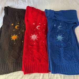 May include: Three sleeveless knit vests in dark brown, bright red, and royal blue. Each vest features a cable knit texture and embroidered celestial motifs including a sun, a crescent moon, and small stars arranged vertically on the front.