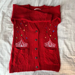 Könnte beinhalten: Roter Zopfstrick-Cardigan mit V-Ausschnitt und Knopfverschluss. Der Pullover hat zwei Vordertaschen und ist mit einem Zirkuszelt-Design sowie bunten Sternen und Punkten bestickt. Die Ärmel sind umgeschlagen.
