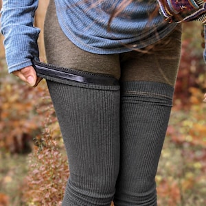 Op de afbeelding: Een blauw shirt met lange geribbelde mouwen en duimgaten en een paar donkergrijze geribbelde beenwarmers. De beenwarmers hebben een zwarte elastische band aan de bovenrand en lopen over de benen, gedragen over een donkerbruine legging in een bosrijke omgeving.