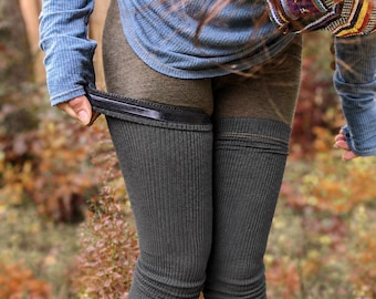 May include: A blue long-sleeved ribbed shirt with thumb holes and a pair of dark gray ribbed leg warmers. The leg warmers feature a black elastic band at the top edge and extend down the legs, worn over dark brown leggings in a forest setting.
