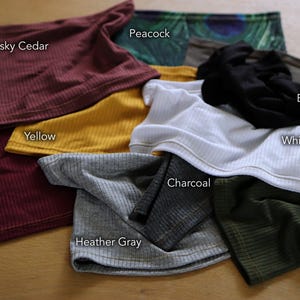 a variety of colorful clothing items, including a red scarf, a black scarf, a white shirt, a maroon scarf, a yellow scarf, a black scarf, a white shirt, a gray scarf, a charcoal scarf, a black scarf, a white shirt, a gray scarf, a black scarf, a white shirt, an olive scarf, and a gray scarf, all laid out on a wooden surface.