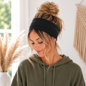 A woman with blonde hair wearing a black headband and a green hoodie is smiling and looking down.