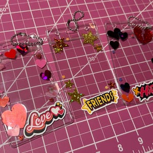 Pode incluir: Tr&ecirc;s molduras de acr&iacute;lico transparente decoradas com cora&ccedil;&otilde;es, estrelas e as palavras "Love", "Friends" e "Hate". Cada moldura tem uma corrente prateada e &eacute; embelezada com glitter e lantejoulas. As molduras est&atilde;o sobre uma superf&iacute;cie de grade rosa.