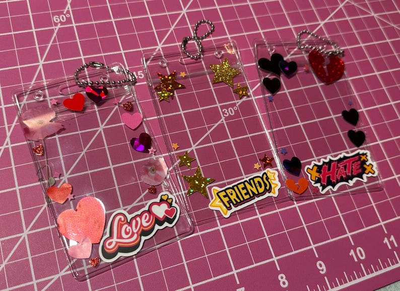 Pode incluir: Tr&ecirc;s molduras de acr&iacute;lico transparente decoradas com cora&ccedil;&otilde;es, estrelas e as palavras "Love", "Friends" e "Hate". Cada moldura tem uma corrente prateada e &eacute; embelezada com glitter e lantejoulas. As molduras est&atilde;o sobre uma superf&iacute;cie de grade rosa.