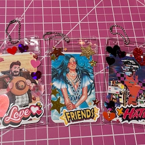 Pode incluir: Tr&ecirc;s porta-crach&aacute;s de pl&aacute;stico transparente com inser&ccedil;&otilde;es decorativas. Um tem uma imagem de cowboy com a palavra "Love", outro uma mulher com a palavra "Friends" e o &uacute;ltimo uma paisagem urbana com a palavra "Hate". Cada um tem enfeites de cora&ccedil;&atilde;o e estrela.