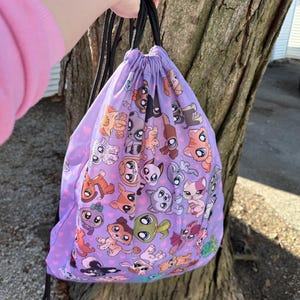 May include: A purple drawstring backpack featuring a repeating pattern of cartoon animals in various colors. The bag has black drawstrings and a pink heart pattern. The bag is held up by a person.