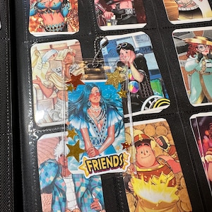 Pode incluir: Uma cole&ccedil;&atilde;o de cartas colecion&aacute;veis e um marcador transparente com uma corrente dourada. O marcador tem uma ilustra&ccedil;&atilde;o de uma mulher de cabelo azul e a palavra "FRIENDS" em uma faixa amarela. As cartas apresentam v&aacute;rias ilustra&ccedil;&otilde;es coloridas de pessoas.