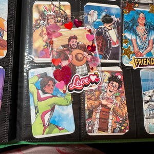 May include: A collection of colorful, illustrated trading cards in protective sleeves. The cards feature various characters, some with decorative embellishments like hearts and the word "Love". The cards are stored in a black binder.