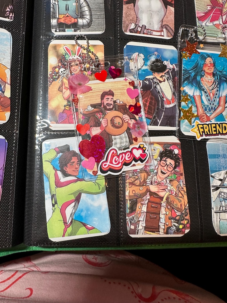 Pode incluir: Uma cole&ccedil;&atilde;o de cartas colecion&aacute;veis ilustradas e coloridas em mangas protetoras. As cartas apresentam v&aacute;rios personagens, alguns com enfeites decorativos como cora&ccedil;&otilde;es e a palavra "Love". As cartas s&atilde;o armazenadas em uma pasta preta.