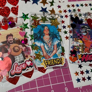 Pode incluir: Tr&ecirc;s chaveiros de acr&iacute;lico transparente com ilustra&ccedil;&otilde;es coloridas e detalhes decorativos. Um apresenta um homem com um chap&eacute;u e a palavra "Love", outro uma mulher com a palavra "Friends", e o &uacute;ltimo com a palavra "Hate". Os chaveiros s&atilde;o adornados com cora&ccedil;&otilde;es e estrelas.