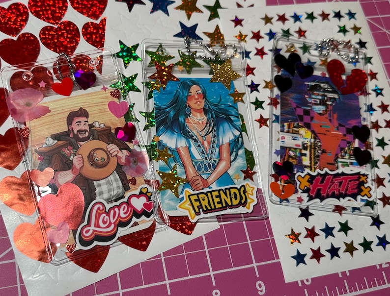 Pode incluir: Tr&ecirc;s chaveiros de acr&iacute;lico transparente com ilustra&ccedil;&otilde;es coloridas e detalhes decorativos. Um apresenta um homem com um chap&eacute;u e a palavra "Love", outro uma mulher com a palavra "Friends", e o &uacute;ltimo com a palavra "Hate". Os chaveiros s&atilde;o adornados com cora&ccedil;&otilde;es e estrelas.
