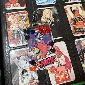 Pode incluir: Um fich&aacute;rio preto contendo cartas colecion&aacute;veis com ilustra&ccedil;&otilde;es em estilo anime. Um pingente de acr&iacute;lico transparente com decora&ccedil;&otilde;es de cora&ccedil;&atilde;o e estrela est&aacute; anexado. Os cart&otilde;es apresentam v&aacute;rios personagens coloridos.