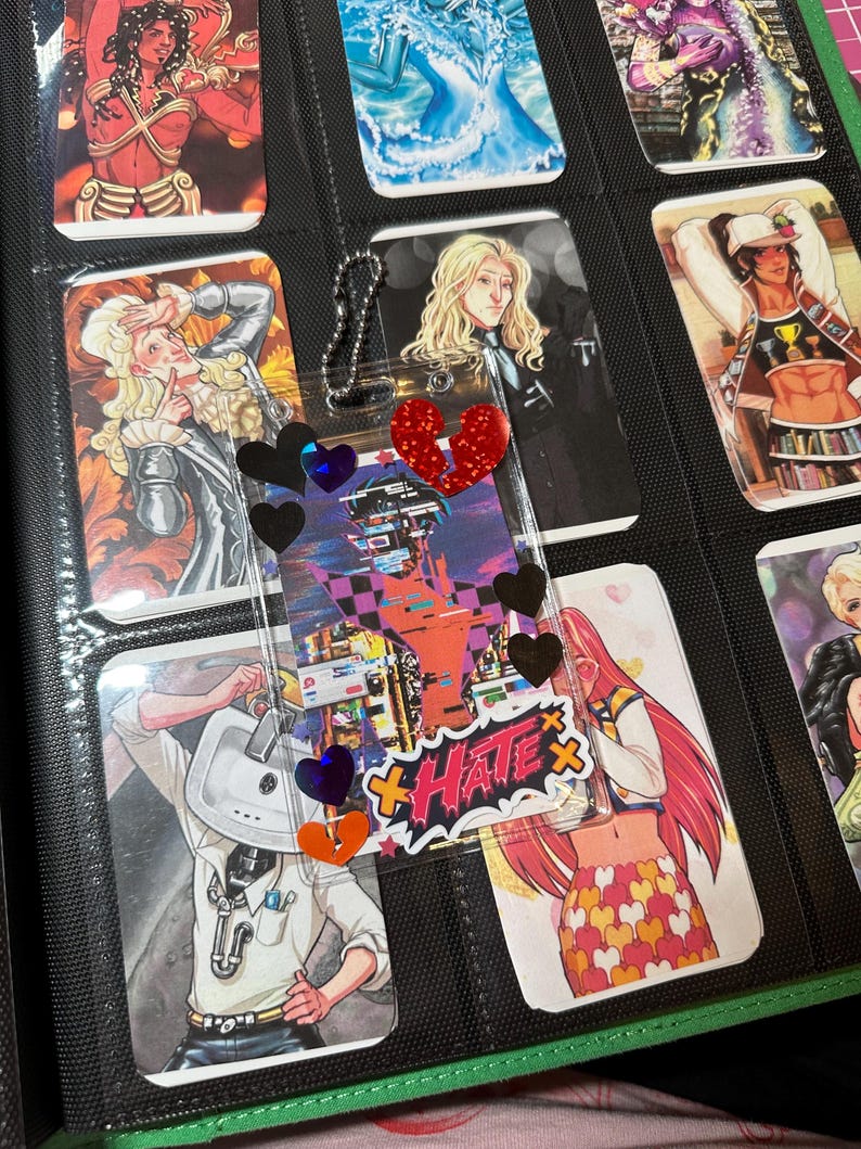 Pode incluir: Um fich&aacute;rio preto contendo cartas colecion&aacute;veis com ilustra&ccedil;&otilde;es em estilo anime. Um pingente de acr&iacute;lico transparente com decora&ccedil;&otilde;es de cora&ccedil;&atilde;o e estrela est&aacute; anexado. Os cart&otilde;es apresentam v&aacute;rios personagens coloridos.