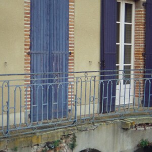 May include: A building with a balcony and two windows with blue shutters. The balcony has a wrought iron railing. The building is made of brick and has a yellow facade.