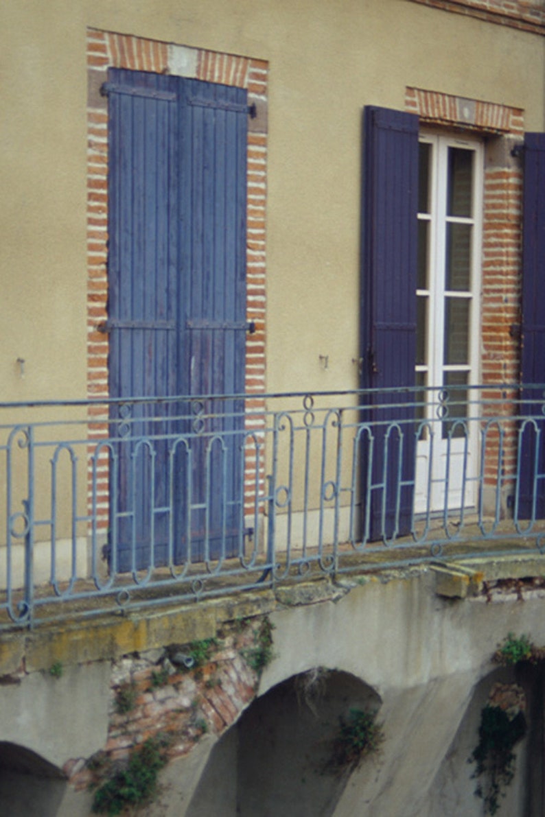 May include: A building with a balcony and two windows with blue shutters. The balcony has a wrought iron railing. The building is made of brick and has a yellow facade.